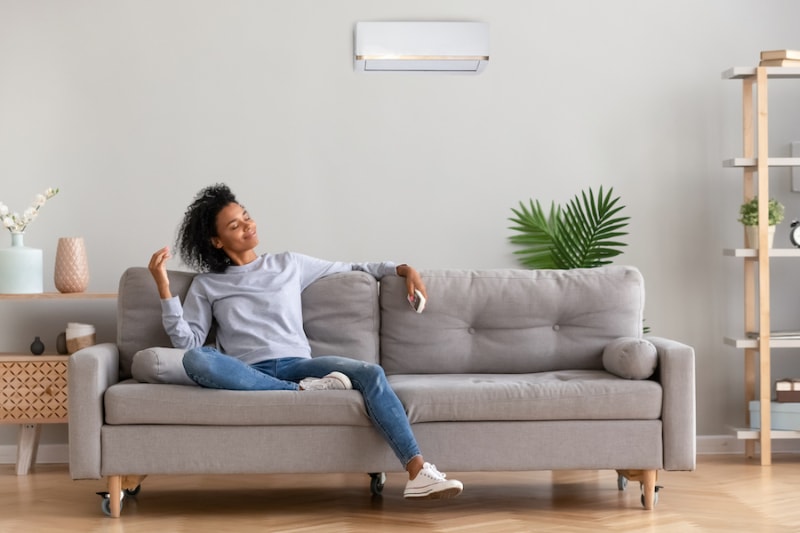 Ductless Mini Splits for Comfortable and Healthy Living. African american relaxed woman sitting on comfortable couch in living room at modern home holds air conditioner remote control enjoying breathing fresh cool air at summer or warm air at winter season African American relaxed woman sitting on comfortable couch.