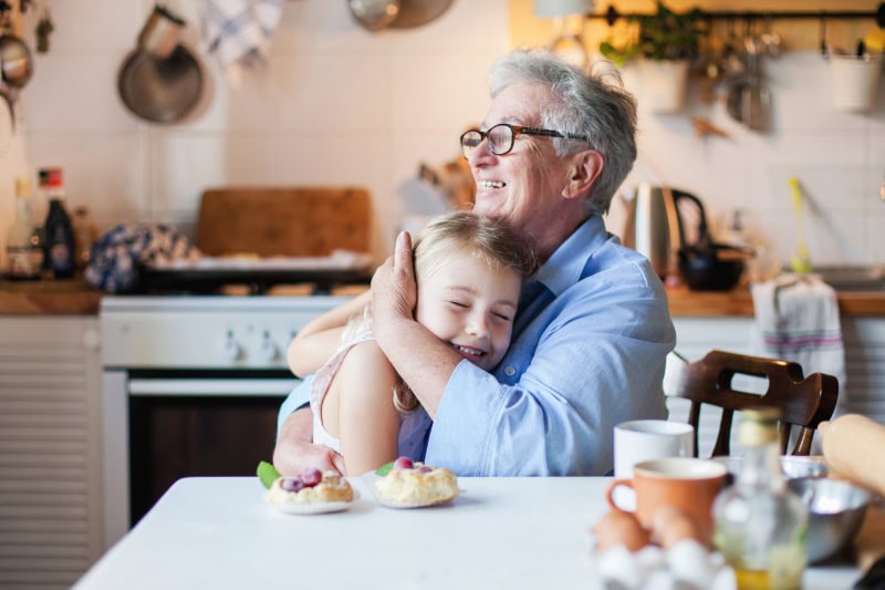 Happy grandmother is hugging granddaughter in cozy home kitchen. Family is cooking together.